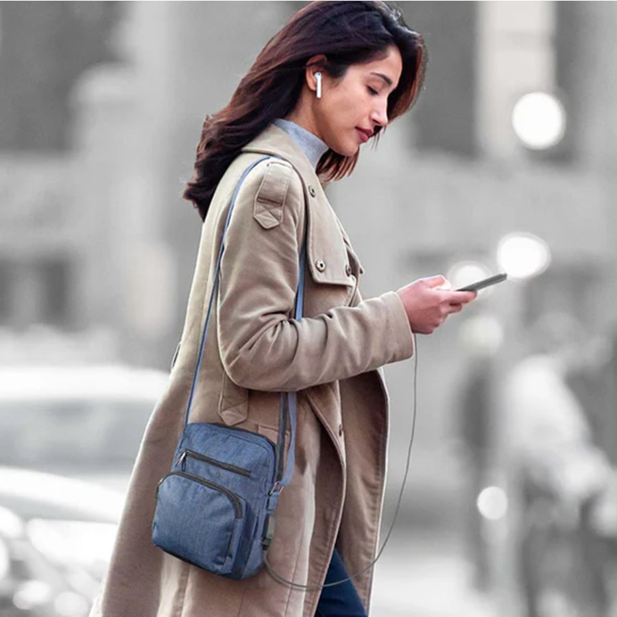 Woman in a beige coat with a blue crossbody bag using a phone on a city street.