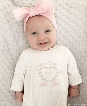 Baby wearing a pink bow headband and white outfit with floral design on a textured gray background