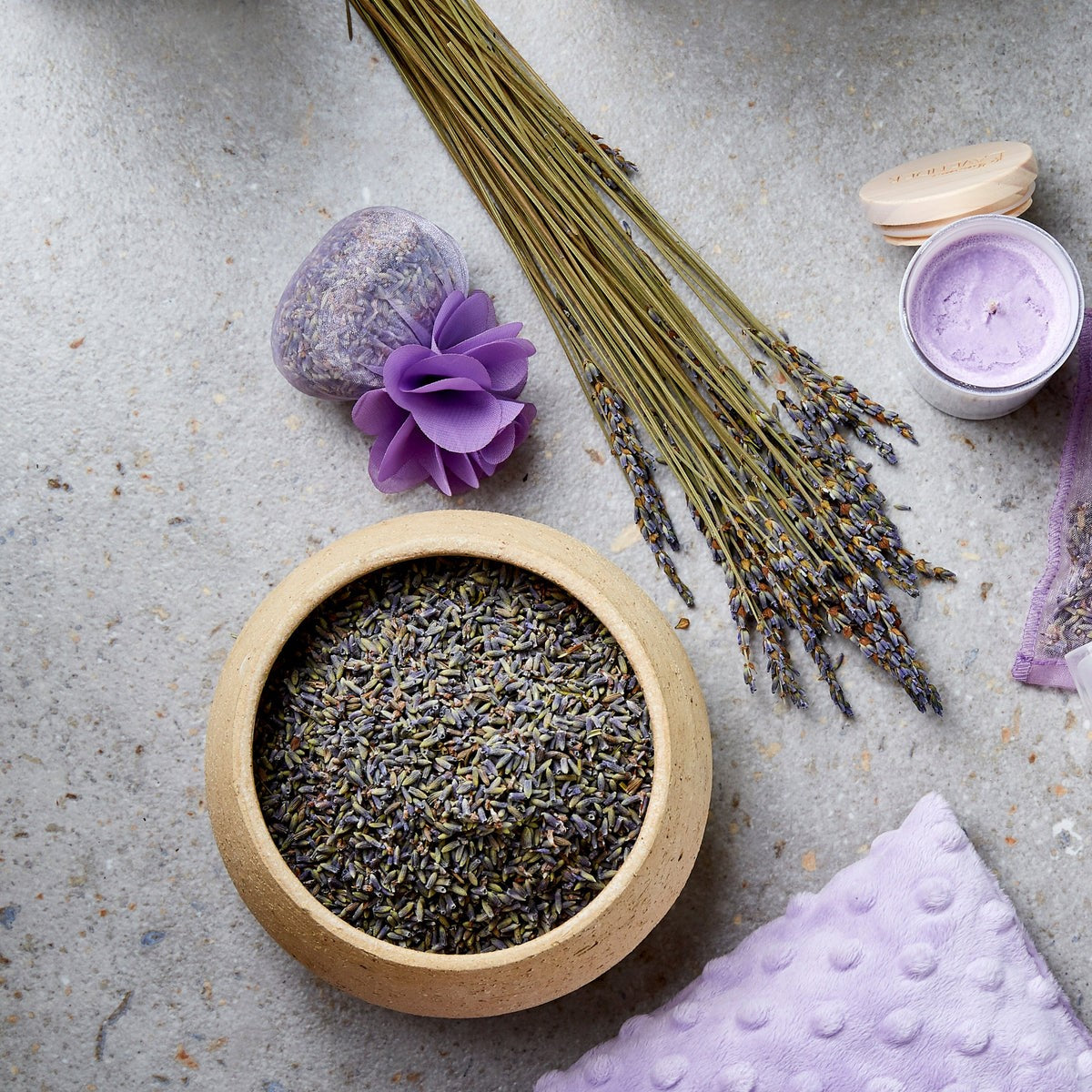 Bowl of dried lavender flowers with a candle, soap, and lavender flower on a stone surface