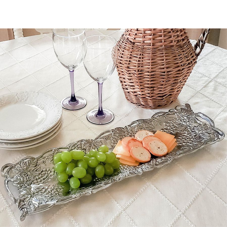 Decorative silver tray with grapes and bread on a table with wine glasses and a basket.