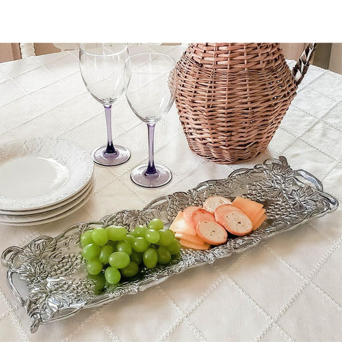 Decorative silver tray with grapes and bread on a table with wine glasses and a basket.