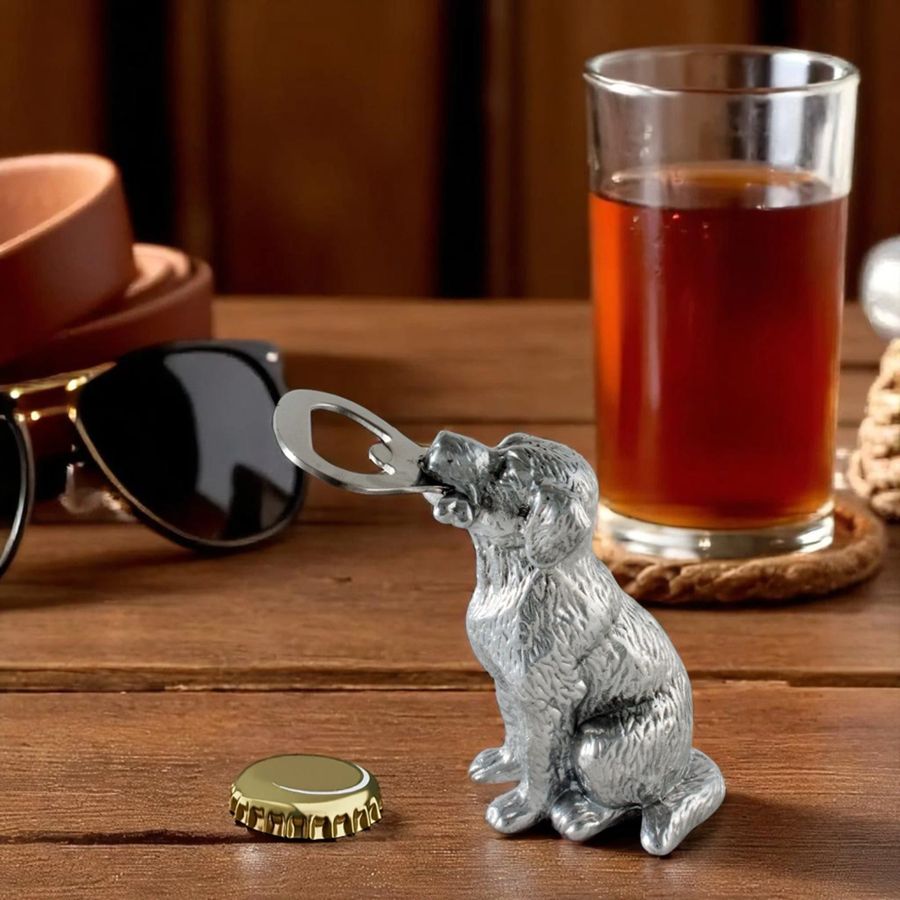Silver dog-shaped bottle opener on a wooden table with a glass of amber liquid and sunglasses in the background.
