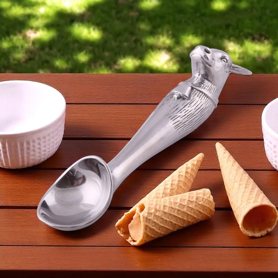 Metal ice cream scoop with cow handle on a wooden surface with waffle cones and bowls.