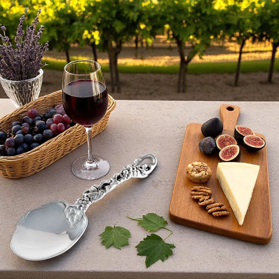 Wine glass with red wine, basket of grapes, cheeseboard with cheese and figs, and silver spoon on a table outdoors.