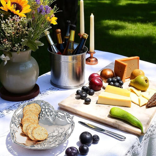 Outdoor setting with a cheeseboard, crackers, fruits, and candles on a tablecloth.