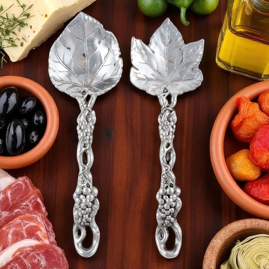 Two silver leaf-shaped spoons on a wooden surface with food items around.