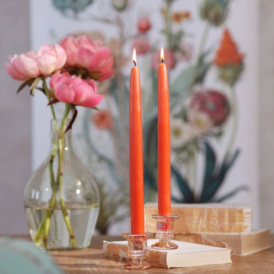 Two lit orange taper candles in glass candlesticks, beside pink flowers in a vase, on books.