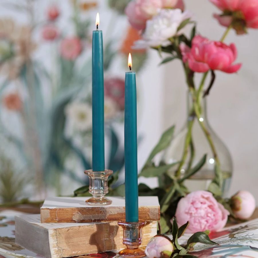 Two blue candles in glass holders on a wooden block with flowers in the background.