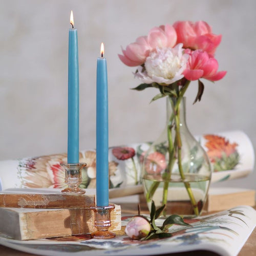 Two blue candles in glass holders on a table with a vase of flowers and a book in the background.