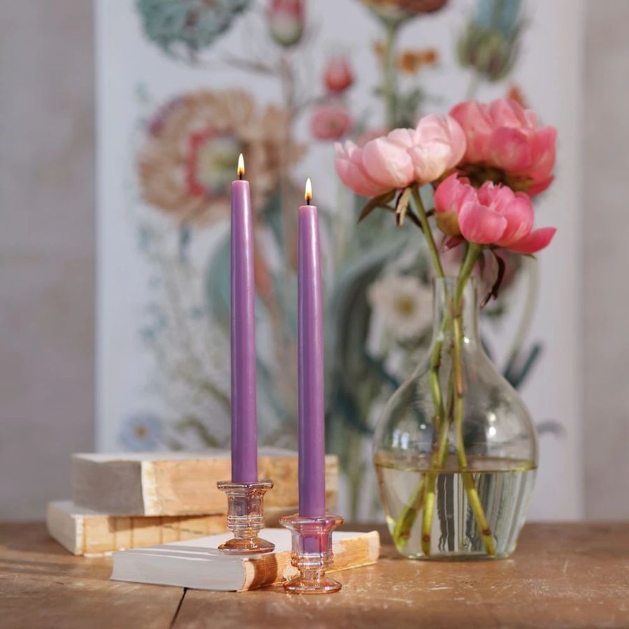 Two purple candles in glass holders on a wooden surface with a vase of pink flowers and a decorative painting in the background.