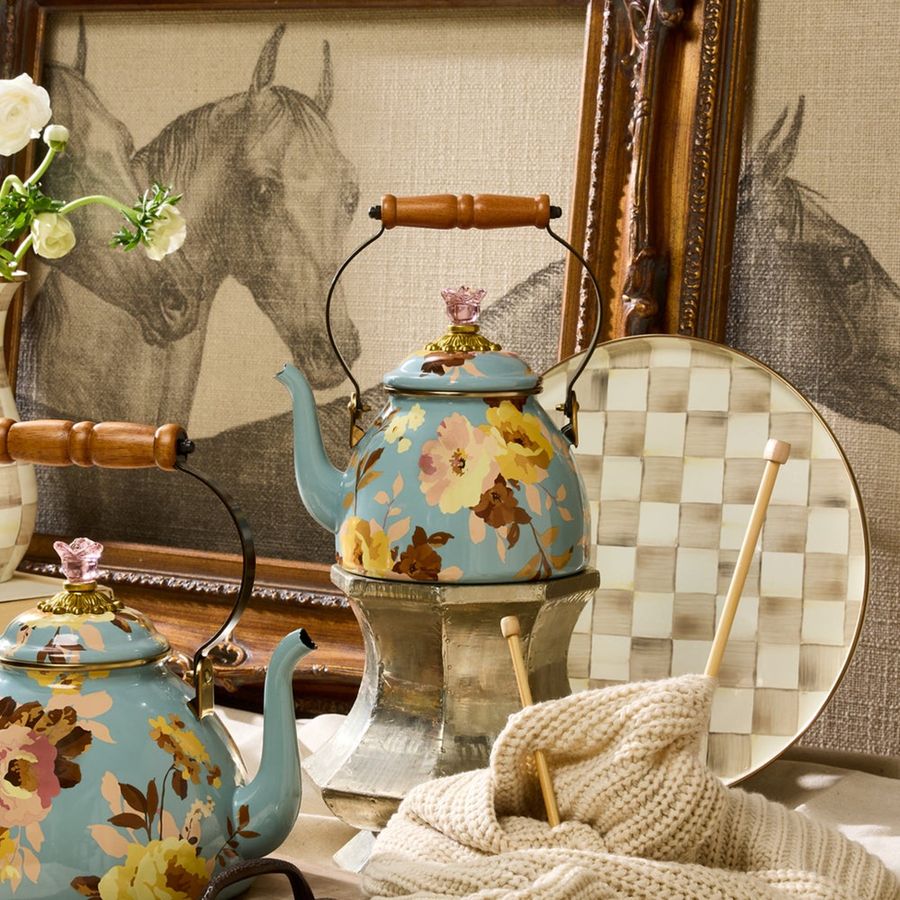 Floral teapot on a table with a checkered cloth and framed horse pictures in the background.