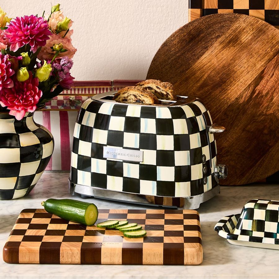 Checkered toaster with bread, cutting board with cucumber, and flowers on a checkered tablecloth.
