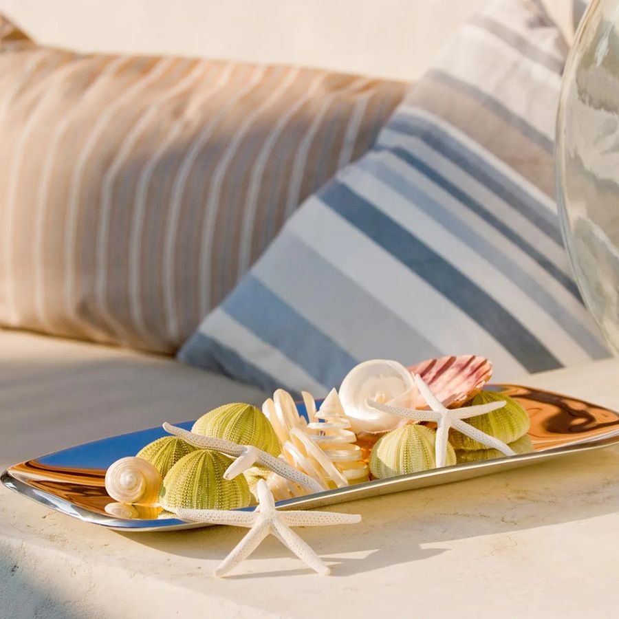 Decorative plate with seashells and starfish on a sandy surface with a blurred chair in the background.