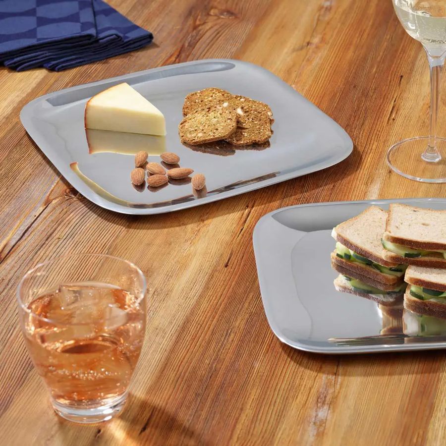 Two square gray trays on a wooden table with food items including bread, butter, almonds, and a glass of juice.