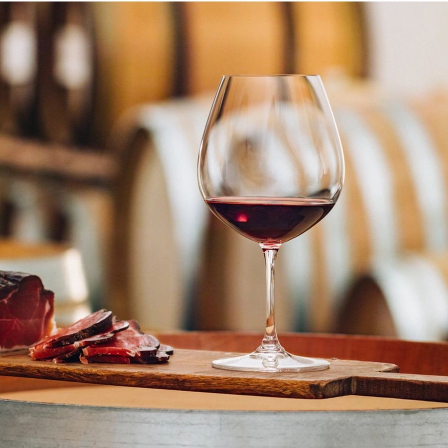 Reidel wine glass with red wine, clear glass, wooden board, sliced cured meat, wine barrels background.