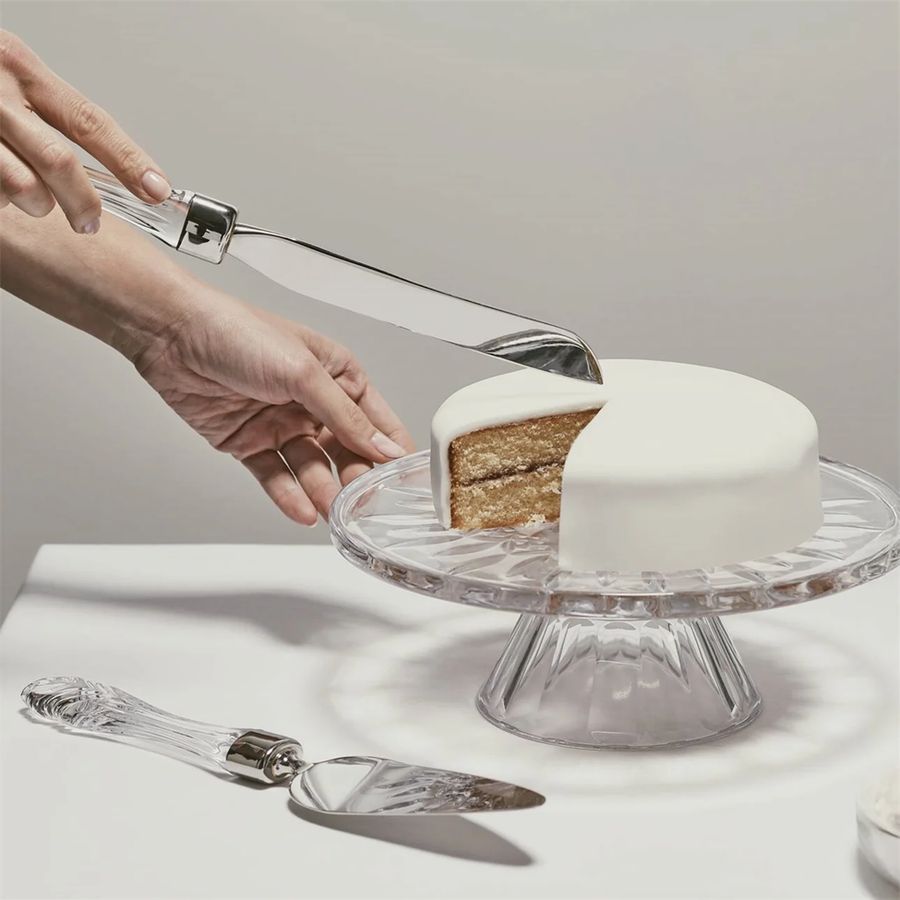Person cutting a white cake on a clear cake stand with a cake knife.