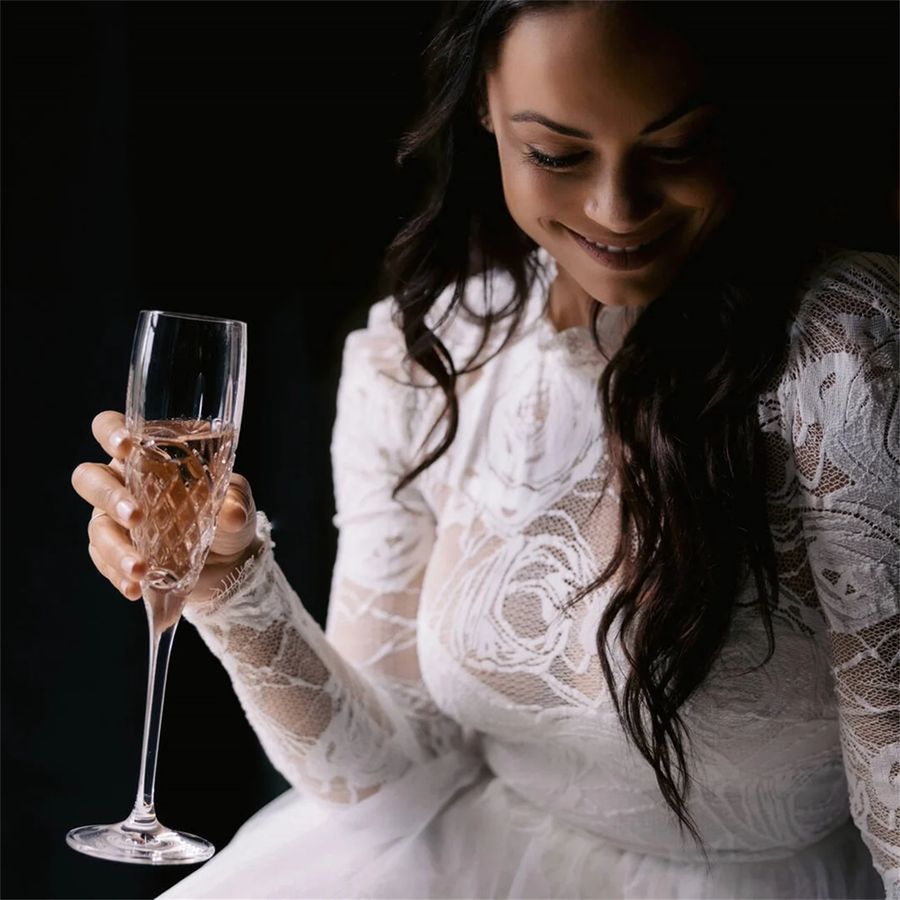 Woman in a white lace dress holding a glass of champagne against a dark background
